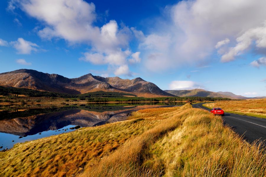 Een rode auto rijdt over een weg dwars door Connemara met uitzicht op de heuvel