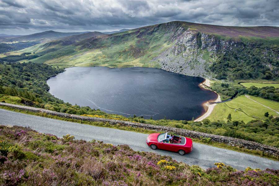 Een rode auto rijdend over een weg door de Glendalough Mountains
